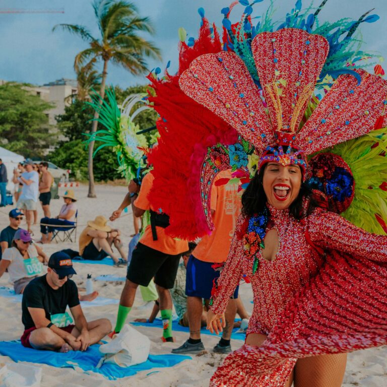 A joyful woman in a vibrant red Carnival costume with large feathered headdress dances on a tropical beach, surrounded by relaxed beachgoers.