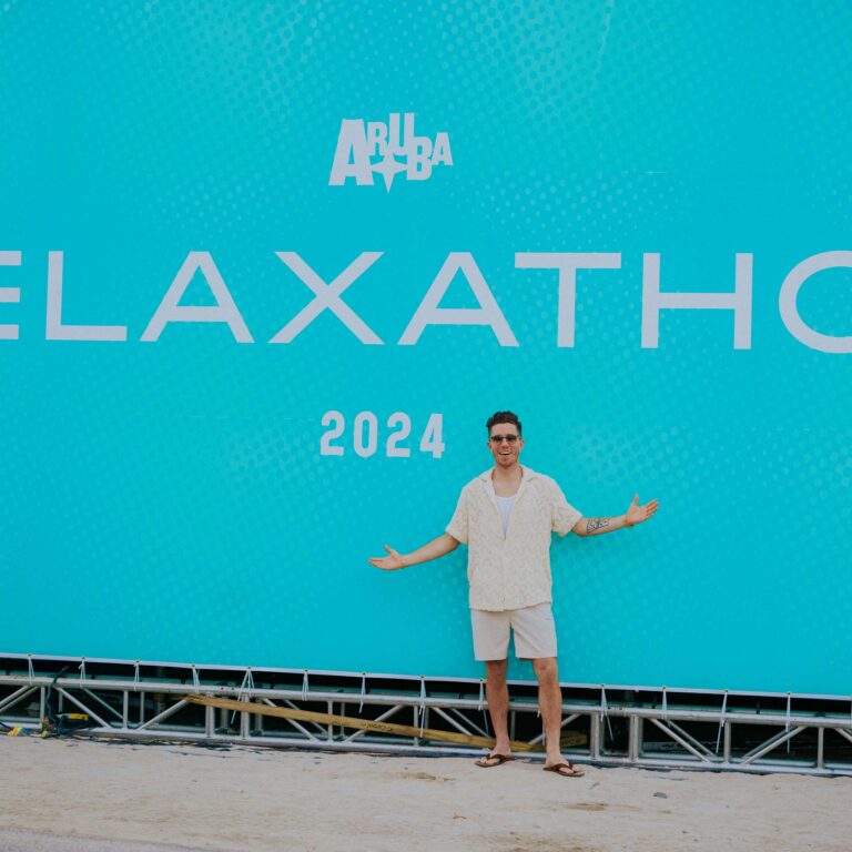 Olympic Snowboarder Shaun White stands smiling with arms open in front of a large turquoise banner that reads "Aruba Relaxathon 2024," conveying a relaxed and joyful mood.