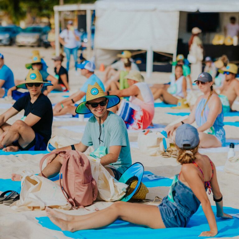 A group of people are sitting on a sandy beach wearing colorful sun hats and casual clothes. They are on bright blue towels, seeming relaxed and enjoying the sunny day.