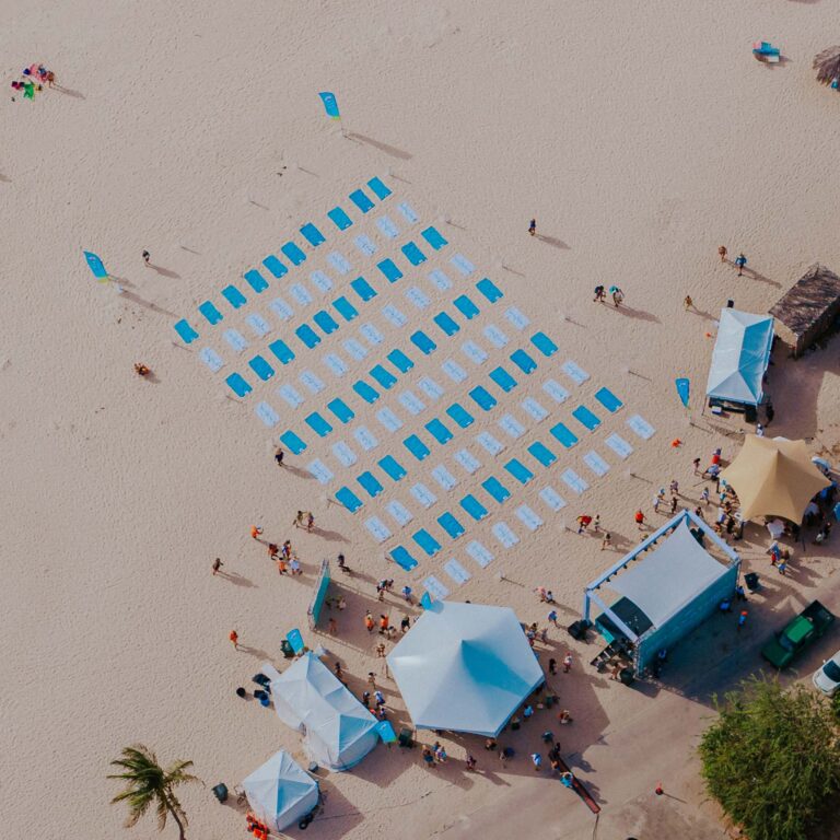 Aerial view of a beach with rows of blue and white towels arranged neatly. People gather near tents and palm trees, creating a lively and organized atmosphere.