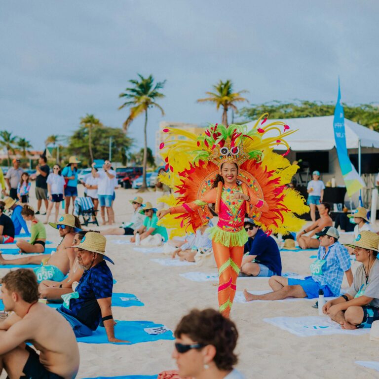 Carnival dancer in a vibrant, feathered costume smiles on a beach, surrounded by seated onlookers. Palm trees and a festive atmosphere in the background.